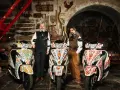 Due uomini in abiti tradizionali posano con tre scooter decorati. Uno scooter non si accende il quadro, ma l'arte sui veicoli è vibrante.