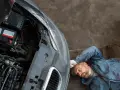 Meccanico con tuta blu accanto a un'auto con cofano aperto, batteria visibile. Pensando a come caricare batteria moto senza caricabatteria.