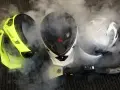 Tre caschi da moto circondati da vapore. Uno giallo brillante, uno bianco e nero, e uno bianco con grafica. Un accessorio per la pulizia a vapore è visibile in primo piano. Il casco deve essere pulito con solventi.