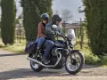 Coppia su una Moto Guzzi V7, con passeggero, percorre una strada alberata in una giornata di sole.