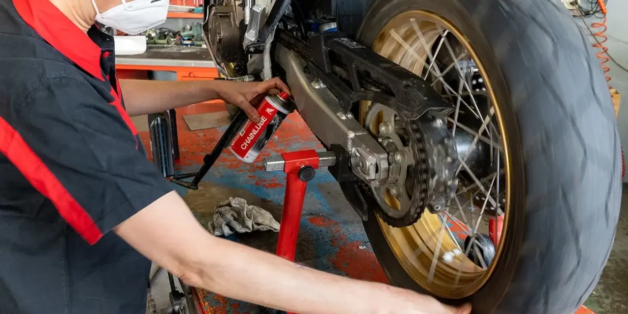 Meccanico con mascherina esegue la pulizia motore e lubrificazione catena moto su cavalletto.