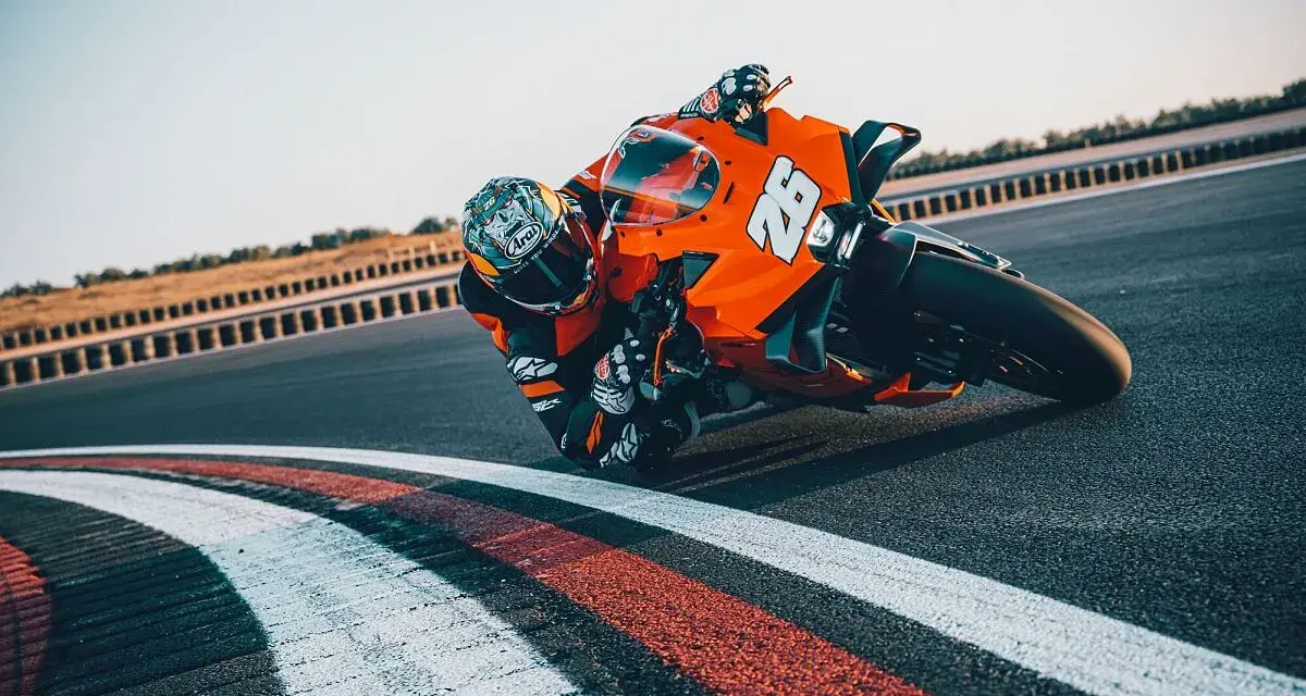 Un pilota con tuta arancione e casco decorato piega in curva su una KTM su una pista da corsa.