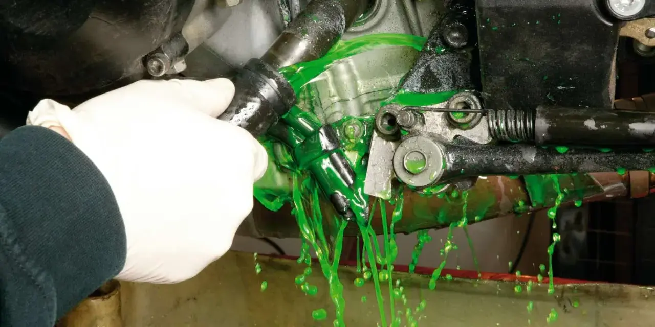 Mano con guanto bianco che scollega un tubo, facendo fuoriuscire liquido verde brillante per il cambio del liquido radiatore.
