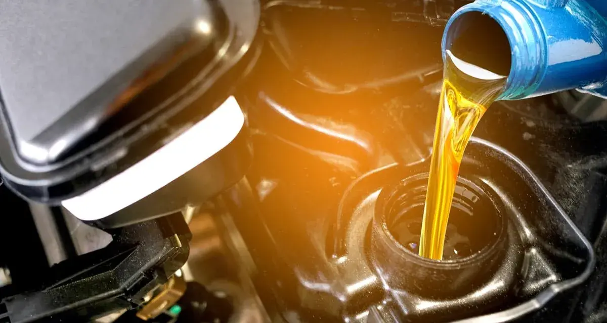 Olio giallo versato da una bottiglia blu in un motore. La differenza tra olio sintetico e minerale è fondamentale per la salute del motore.