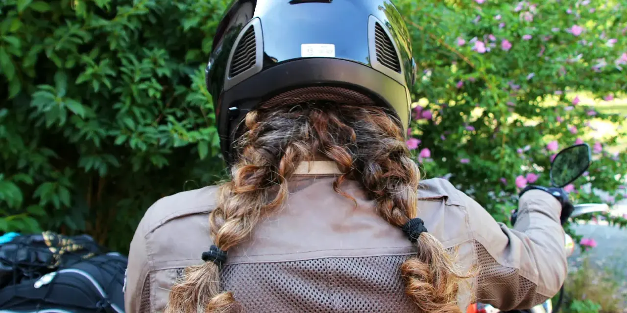 Donna con casco nero e capelli raccolti in due trecce, pronta a legare il casco alla moto.