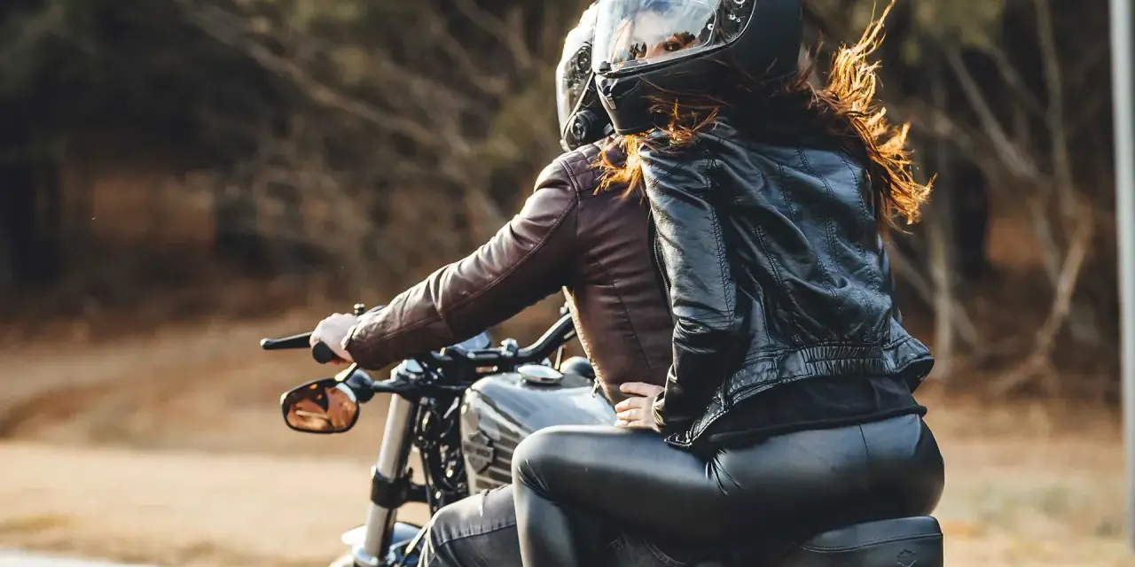 Coppia in moto, lei indossa giacca di pelle e pantaloni neri, lui giacca marrone. Perfetto esempio di come vestirsi per andare in moto passeggero.