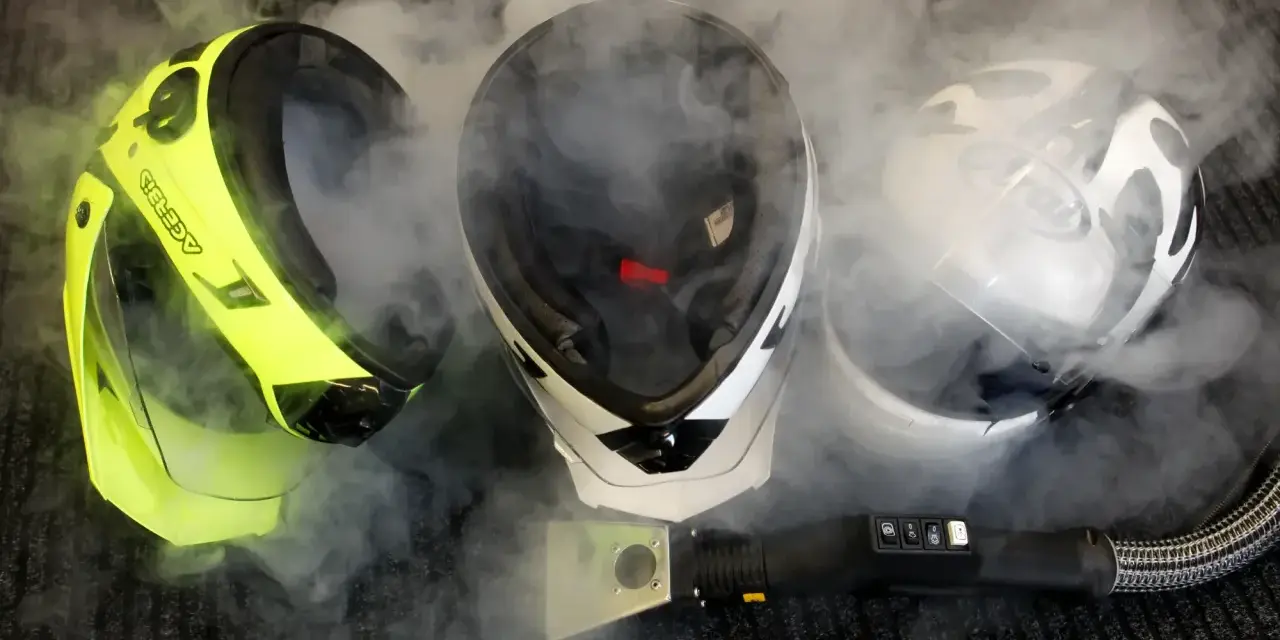 Tre caschi da moto circondati da vapore. Uno giallo brillante, uno bianco e nero, e uno bianco con grafica. Un accessorio per la pulizia a vapore è visibile in primo piano. Il casco deve essere pulito con solventi.