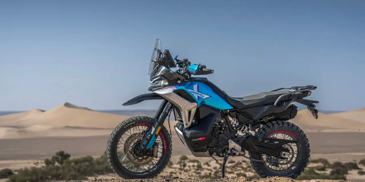 Moto blu e nera, perfetta per avventura e viaggi, posizionata su un terreno sabbioso con dune all'orizzonte.