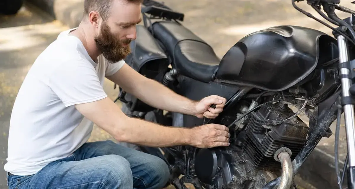 Uomo con barba lavora sul motore di una moto, forse per sostituire subito la batteria.