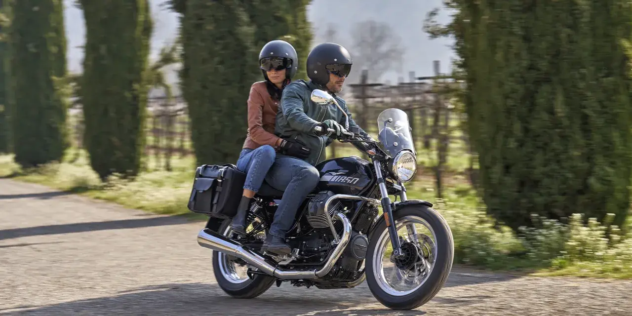 Coppia su una Moto Guzzi V7, con passeggero, percorre una strada alberata in una giornata di sole.