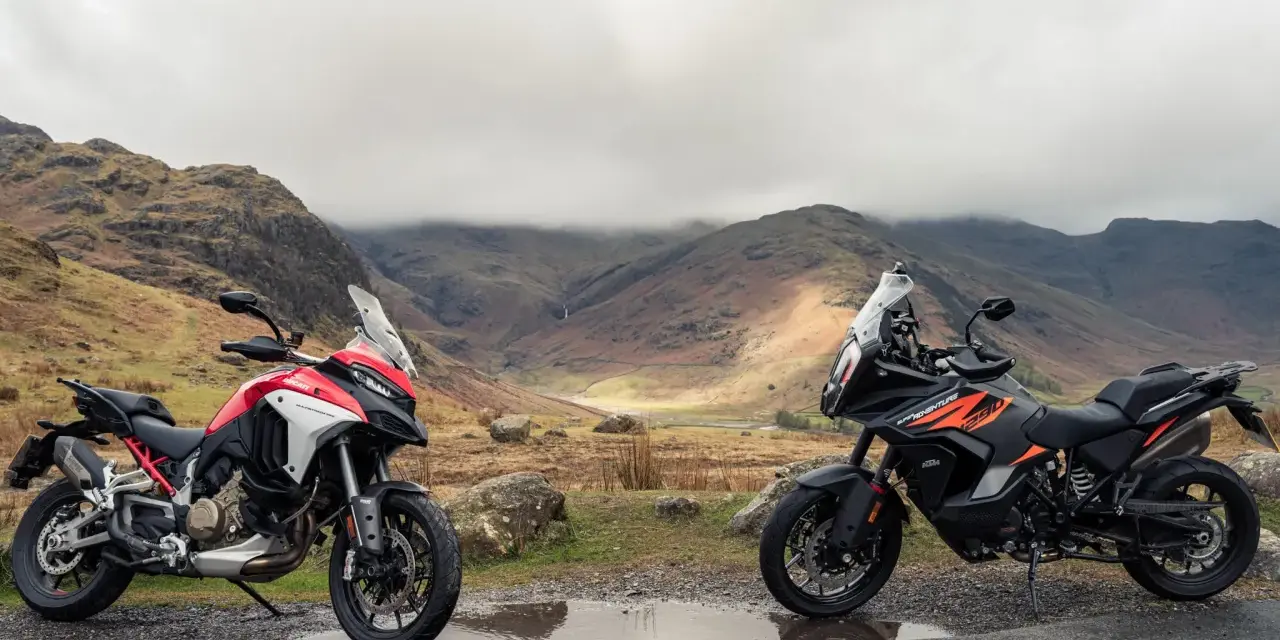 Due moto, una Ducati Multistrada 1290 rossa e bianca e una KTM 1290 Super Adventure nera e arancione, parcheggiate su una strada di montagna con un paesaggio nebbioso sullo sfondo.