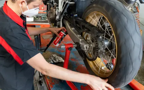 Meccanico con mascherina esegue la pulizia motore e lubrificazione catena moto su cavalletto.