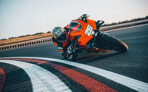 Un pilota con tuta arancione e casco decorato piega in curva su una KTM su una pista da corsa.