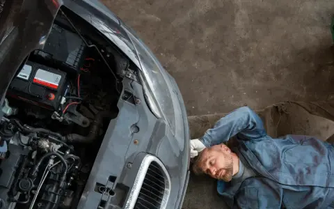 Meccanico con tuta blu accanto a un'auto con cofano aperto, batteria visibile. Pensando a come caricare batteria moto senza caricabatteria.