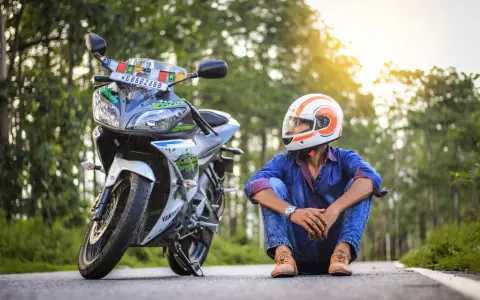 Giovane con casco da moto arancione e bianco, seduto accanto alla sua Yamaha. Spera di allargare il casco stretto per un viaggio più comodo.