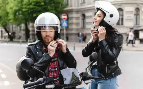 Coppia in moto, entrambi indossano un casco integrale. Lui con camicia a quadri, lei con giacca di pelle.