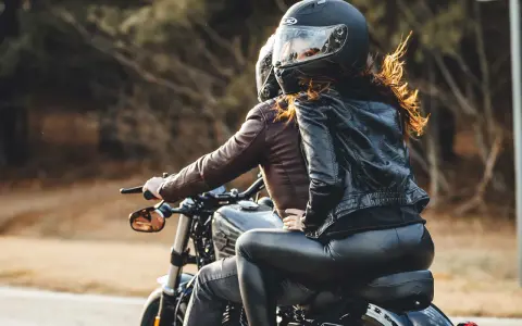 Coppia in moto, lei indossa giacca di pelle e pantaloni neri, lui giacca marrone. Perfetto esempio di come vestirsi per andare in moto passeggero.