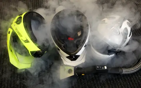 Tre caschi da moto circondati da vapore. Uno giallo brillante, uno bianco e nero, e uno bianco con grafica. Un accessorio per la pulizia a vapore è visibile in primo piano. Il casco deve essere pulito con solventi.