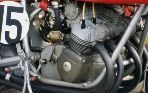 Dettaglio del motore di una moto da corsa, con il numero 15 su un cerchio bianco. Il motore è un complesso di tubi e metallo, pronto a ruggire.