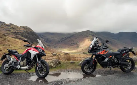 Due moto, una Ducati Multistrada 1290 rossa e bianca e una KTM 1290 Super Adventure nera e arancione, parcheggiate su una strada di montagna con un paesaggio nebbioso sullo sfondo.
