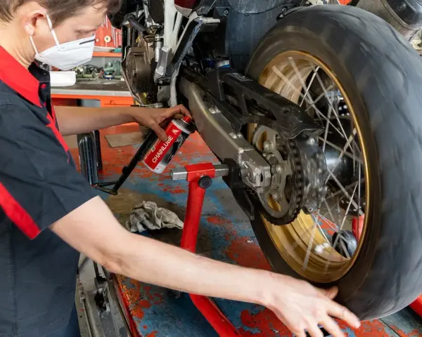 Meccanico con mascherina esegue la pulizia motore e lubrificazione catena moto su cavalletto.