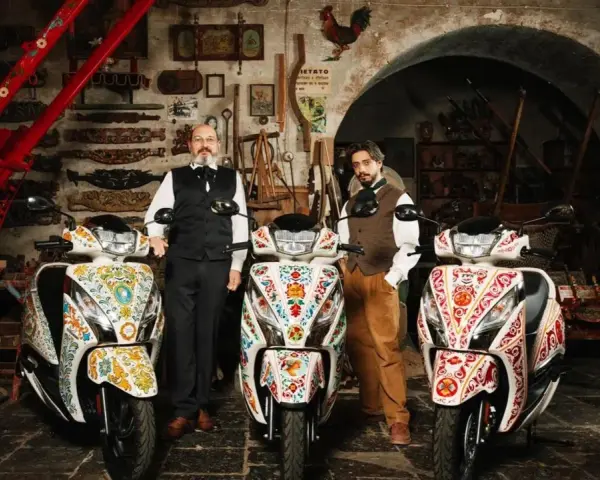 Due uomini in abiti tradizionali posano con tre scooter decorati. Uno scooter non si accende il quadro, ma l'arte sui veicoli è vibrante.