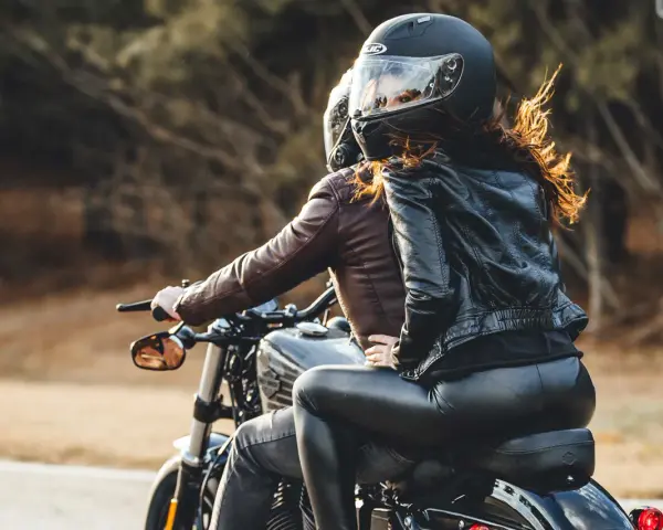 Coppia in moto, lei indossa giacca di pelle e pantaloni neri, lui giacca marrone. Perfetto esempio di come vestirsi per andare in moto passeggero.