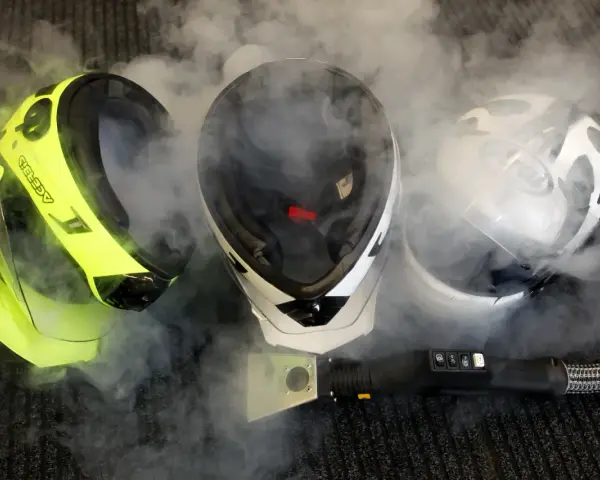 Tre caschi da moto circondati da vapore. Uno giallo brillante, uno bianco e nero, e uno bianco con grafica. Un accessorio per la pulizia a vapore è visibile in primo piano. Il casco deve essere pulito con solventi.