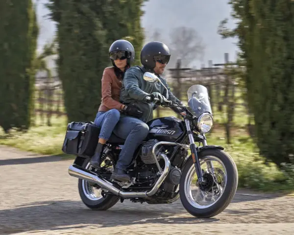 Coppia su una Moto Guzzi V7, con passeggero, percorre una strada alberata in una giornata di sole.