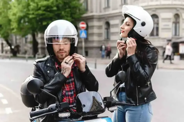 Coppia in moto, entrambi indossano un casco integrale. Lui con camicia a quadri, lei con giacca di pelle.