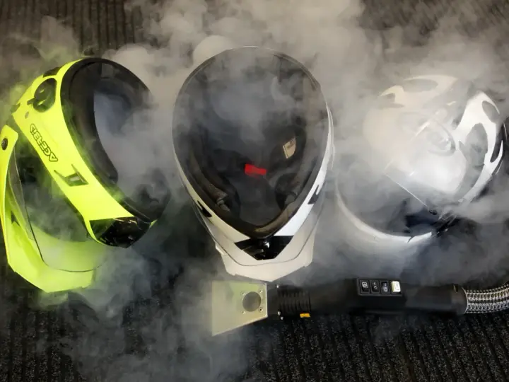 Tre caschi da moto circondati da vapore. Uno giallo brillante, uno bianco e nero, e uno bianco con grafica. Un accessorio per la pulizia a vapore è visibile in primo piano. Il casco deve essere pulito con solventi.