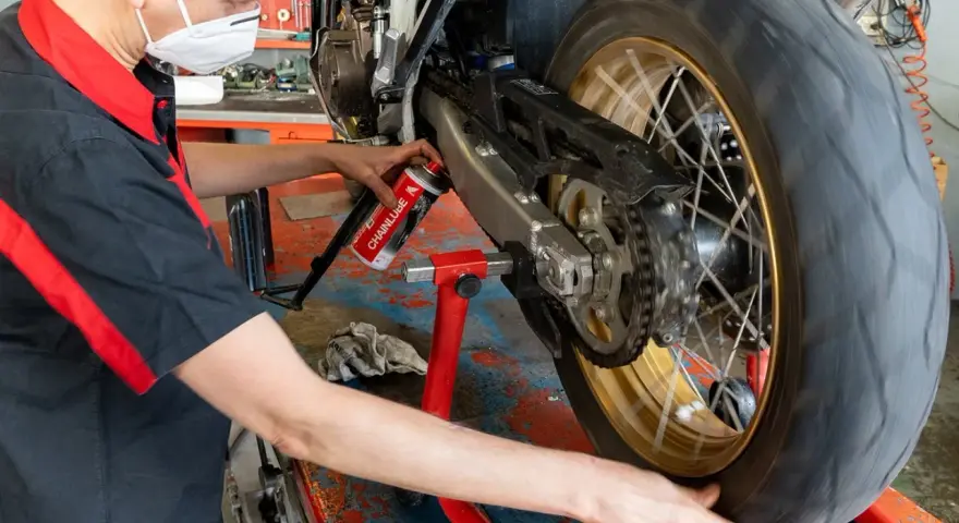 Meccanico con mascherina esegue la pulizia motore e lubrificazione catena moto su cavalletto.