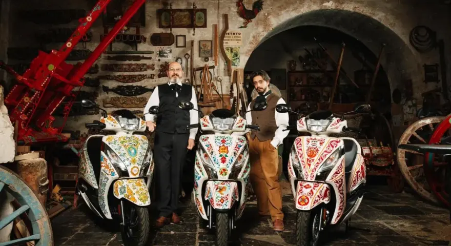 Due uomini in abiti tradizionali posano con tre scooter decorati. Uno scooter non si accende il quadro, ma l'arte sui veicoli è vibrante.