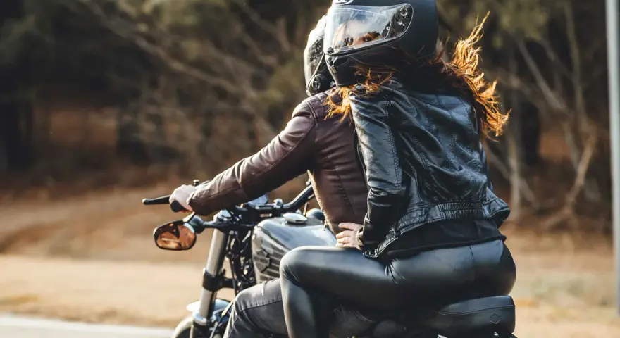 Coppia in moto, lei indossa giacca di pelle e pantaloni neri, lui giacca marrone. Perfetto esempio di come vestirsi per andare in moto passeggero.