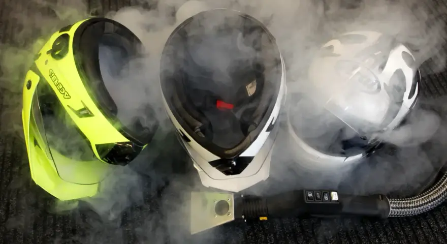 Tre caschi da moto circondati da vapore. Uno giallo brillante, uno bianco e nero, e uno bianco con grafica. Un accessorio per la pulizia a vapore è visibile in primo piano. Il casco deve essere pulito con solventi.