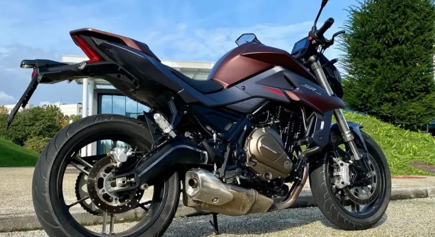 Moto SRK 700 color bronzo e nero, parcheggiata all'aperto con cielo azzurro e verde.