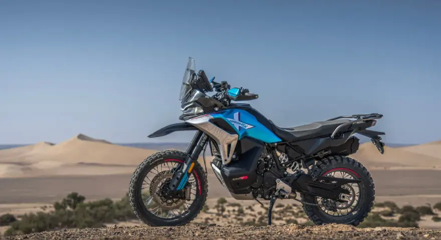 Moto blu e nera, perfetta per avventura e viaggi, posizionata su un terreno sabbioso con dune all'orizzonte.