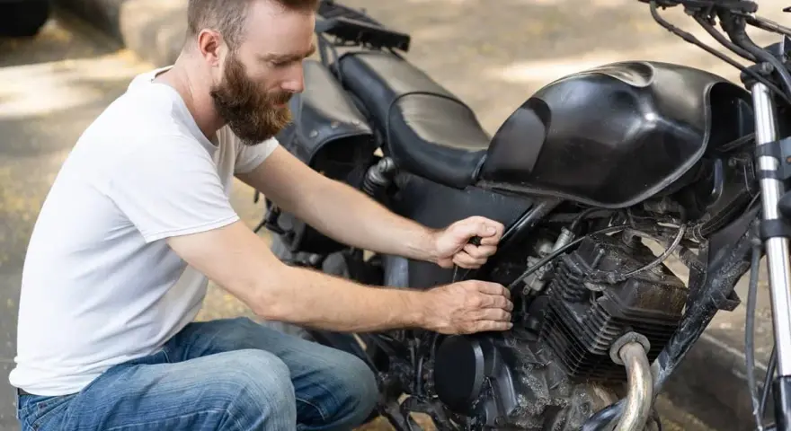 Uomo con barba lavora sul motore di una moto, forse per sostituire subito la batteria.