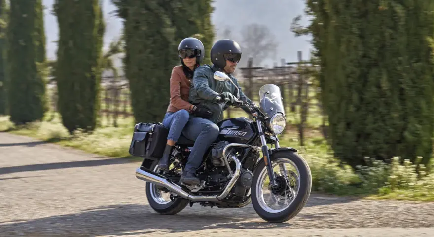 Coppia su una Moto Guzzi V7, con passeggero, percorre una strada alberata in una giornata di sole.