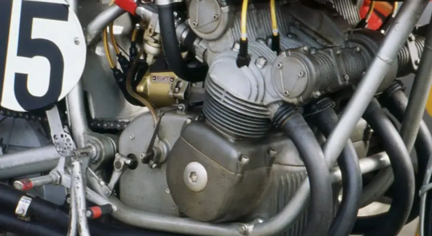 Dettaglio del motore di una moto da corsa, con il numero 15 su un cerchio bianco. Il motore è un complesso di tubi e metallo, pronto a ruggire.