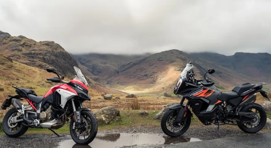 Due moto, una Ducati Multistrada 1290 rossa e bianca e una KTM 1290 Super Adventure nera e arancione, parcheggiate su una strada di montagna con un paesaggio nebbioso sullo sfondo.