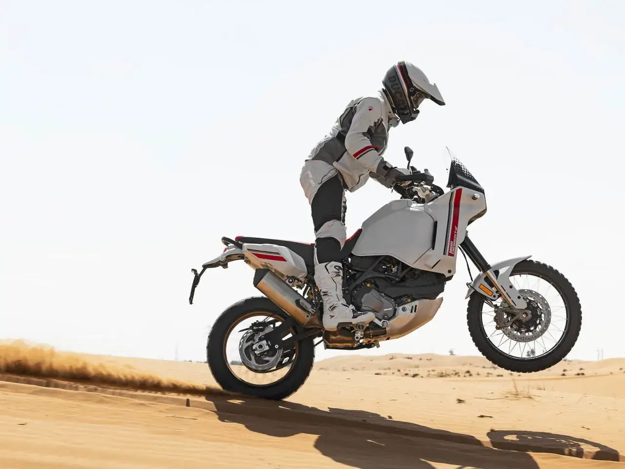 Un pilota in abbigliamento tecnico salta con una moto da enduro bianca nel deserto, sollevando sabbia. L'enduro, un'avventura su due ruote.
