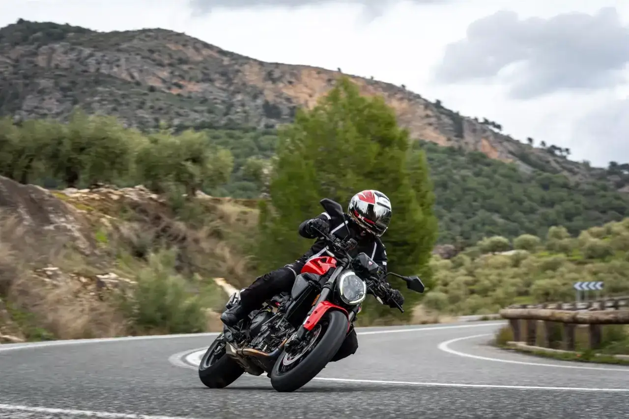 Ducati Monster in curva su strada panoramica, con montagne e alberi sullo sfondo.