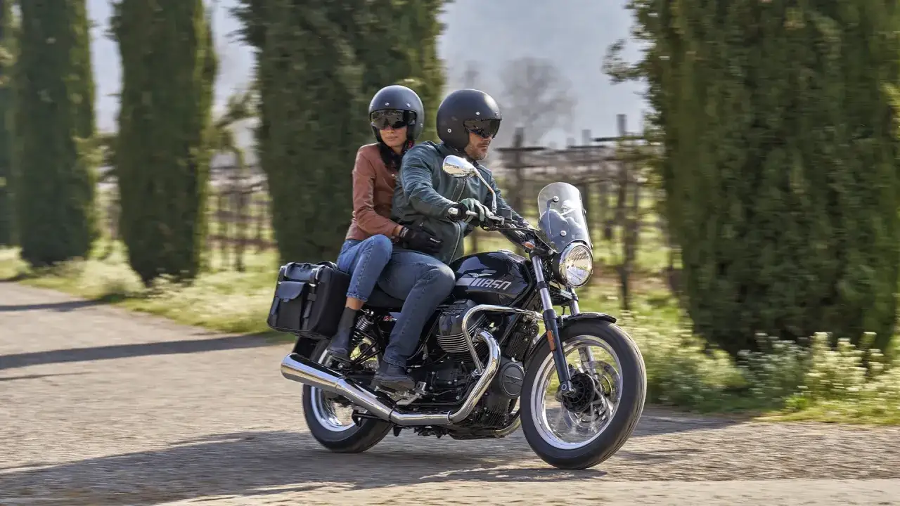 Coppia su una Moto Guzzi V7, con passeggero, percorre una strada alberata.
