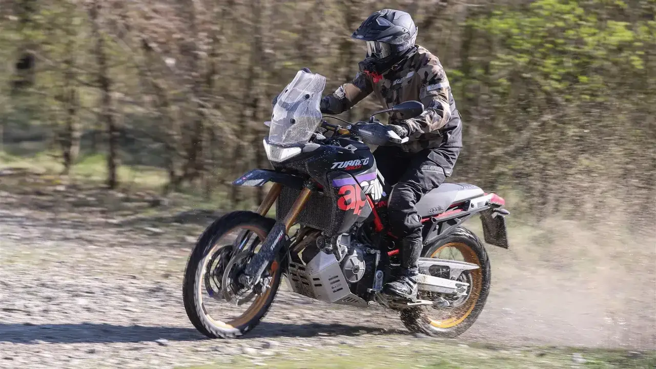 Un pilota su una Aprilia 600 Enduro affronta un percorso sterrato tra gli alberi, sollevando polvere.