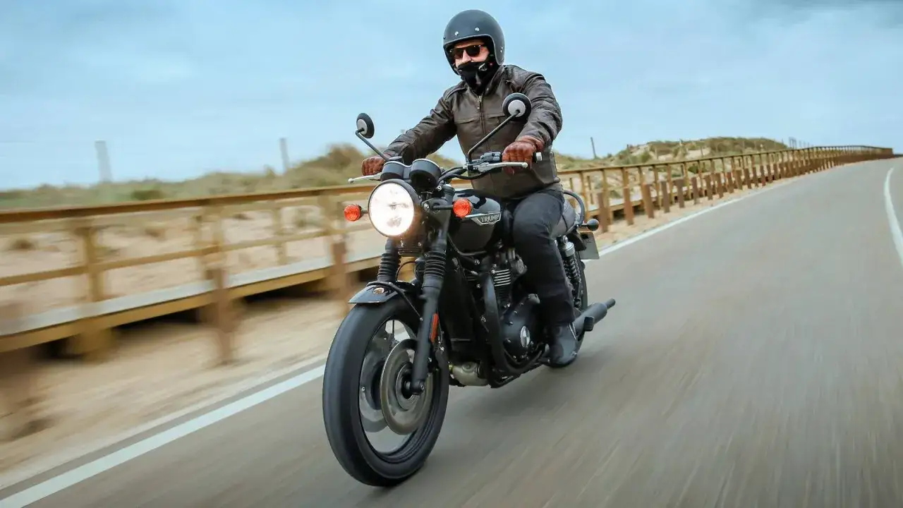 Un motociclista su una moto modern classic sfreccia su una strada costiera, con dune e passerella in legno sullo sfondo.