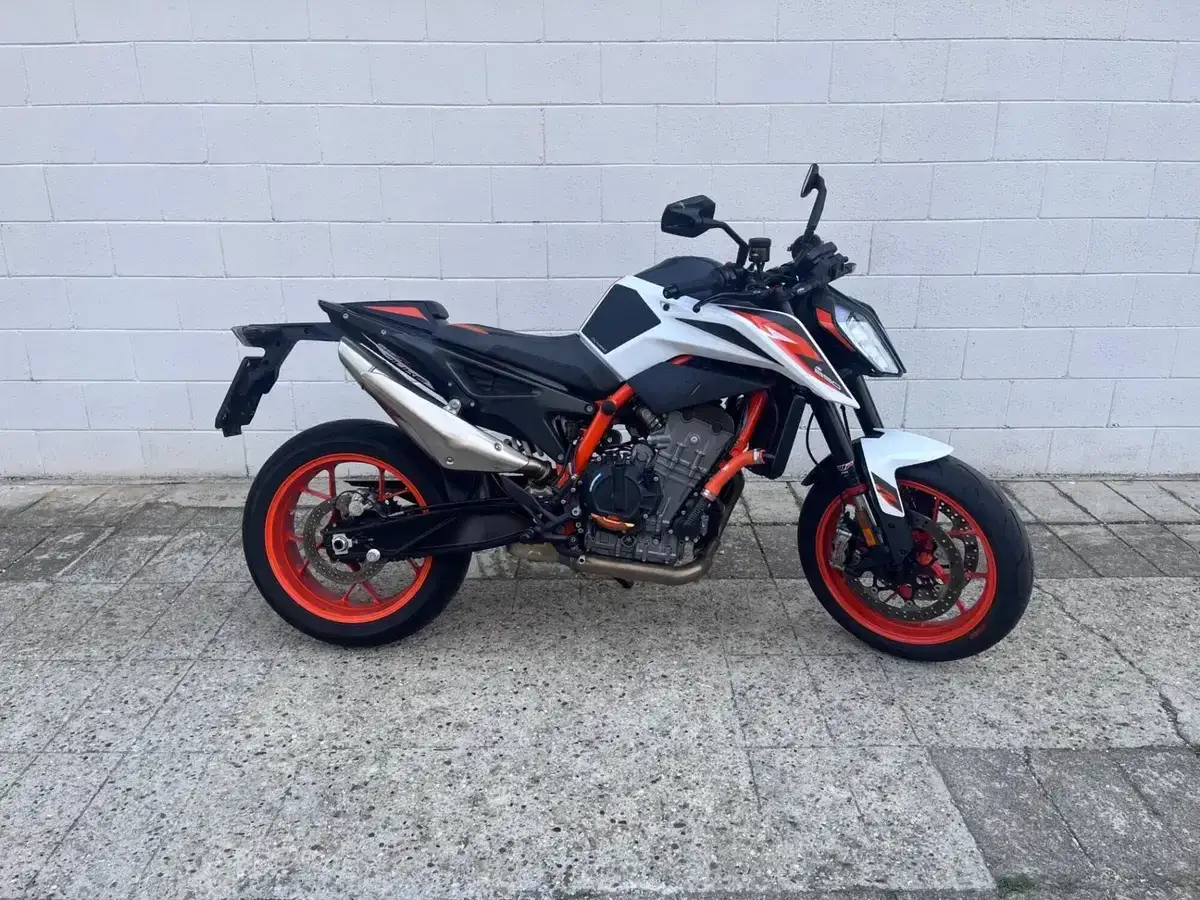 KTM 890 Duke, una moto sportiva bianca e arancione parcheggiata su un marciapiede di cemento davanti a un muro di mattoni bianchi.