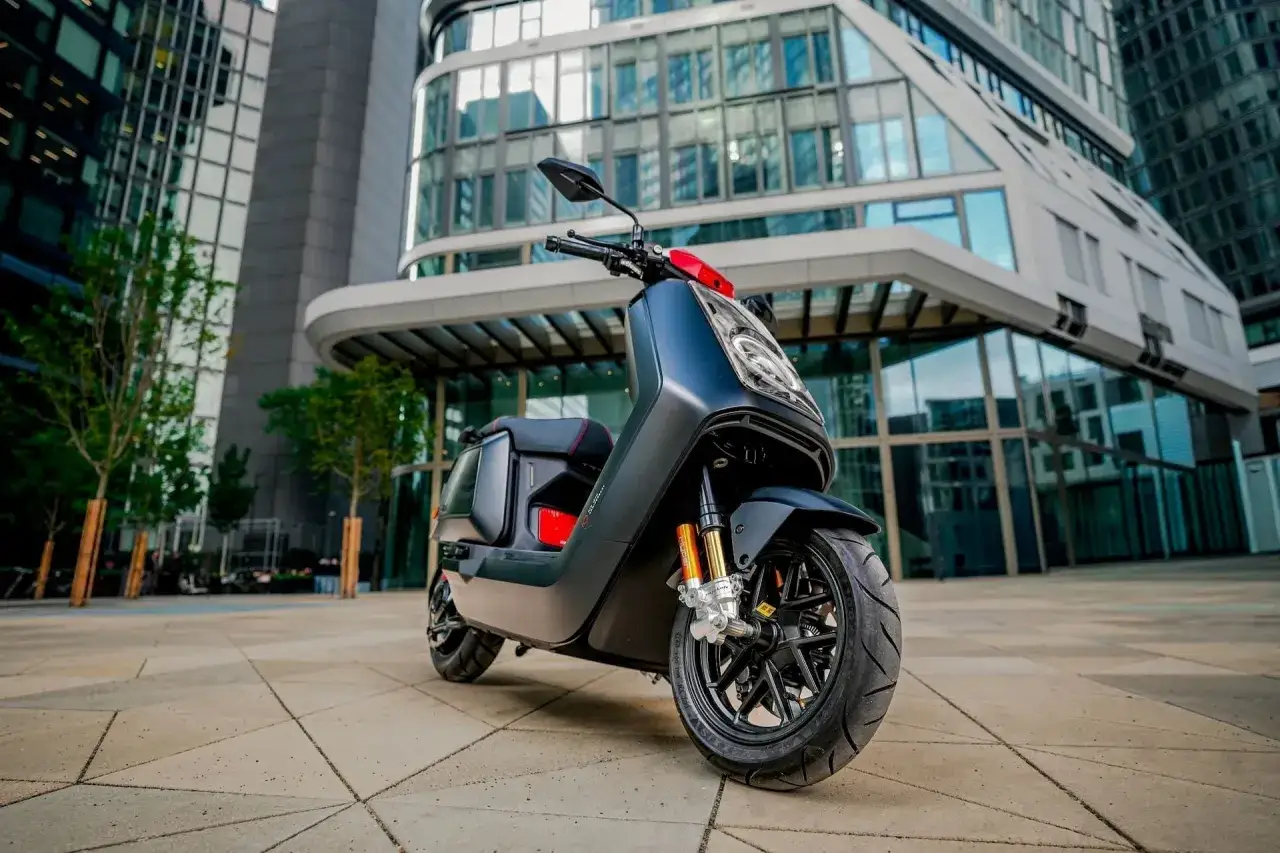 Uno scooter elettrico dal design moderno, con dettagli particolari come le sospensioni arancioni, parcheggiato davanti a un edificio di vetro.