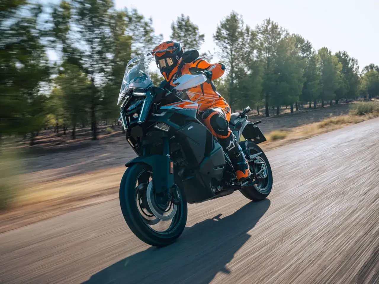 Un motociclista con abbigliamento arancione sfreccia su una KTM maxi enduro su una strada circondata da alberi.