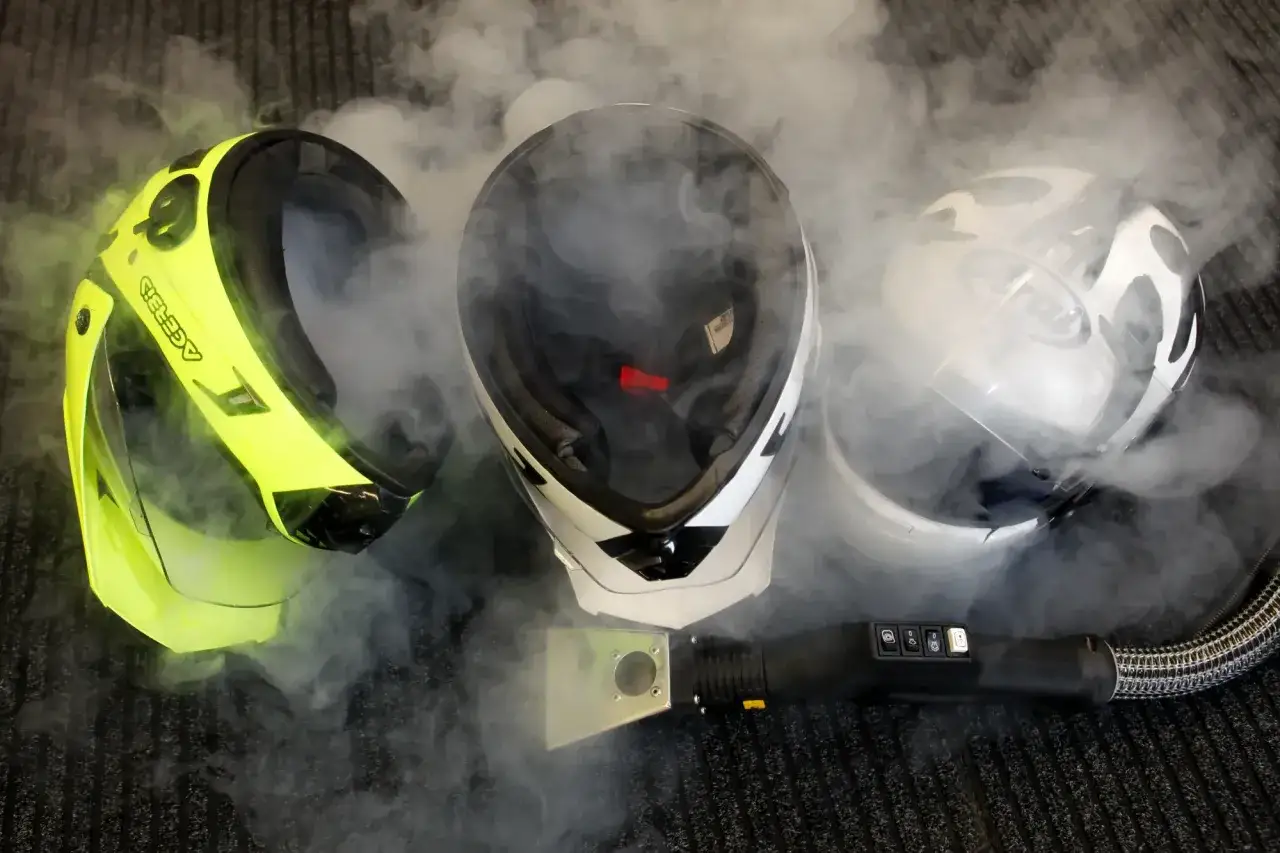 Tre caschi da moto circondati da vapore. Uno giallo brillante, uno bianco e nero, e uno bianco con grafica. Un accessorio per la pulizia a vapore è visibile in primo piano. Il casco deve essere pulito con solventi.